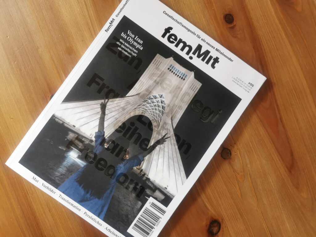 Auf dem Foto ist das femMit-Magazin Ausgabe 5 zu sehen. Es liegt auf einem holzfarbenen Hintergrund. Auf dem Cover des Magazins ist eine Frau zu sehen, die sich an der Revolution im Iran beteiligt.